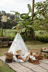 Beautiful mother is hugging and reading a book to her little daughter. Picnic in the park. Wigwam...