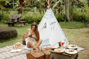 Beautiful girl on the background of the wigwam drinks coffee. Picnic on the green lawn. Outdoor recreation © Yevhenii