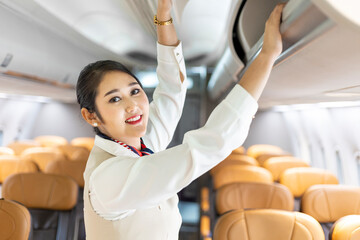 Asian female flight attendant closing the overhead luggage compartment lid for carry on baggage...