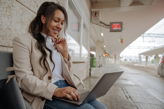 Confident smiling businesswoman using laptop computer having video call sitting on bench. Happy passenger working online waiting for train at railway station. Business trip concept   