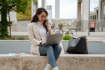 Confident smiling business woman taking on mobile phone, using laptop computer working outdoors. Young freelancer copywriter holding smartphone, typing on keyboard at workplace. Successful business 