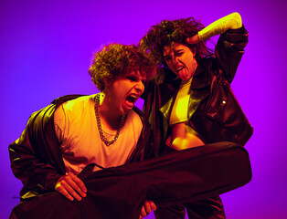 Young couple of rock-and-roll musicians wearing black leather outfits gesturing, shouting and posing on blue yellow background in neon light