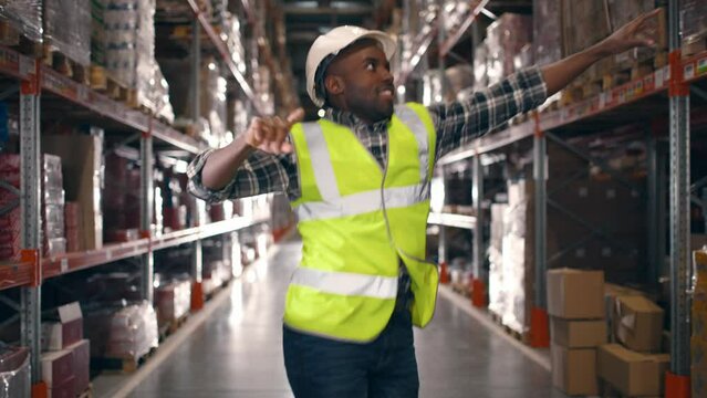 Cheerful African American young male worker in hard hat jumping and dancing between shelves with boxes and goods. Construction, factory, logistics center, work, positive emotions, job concept
