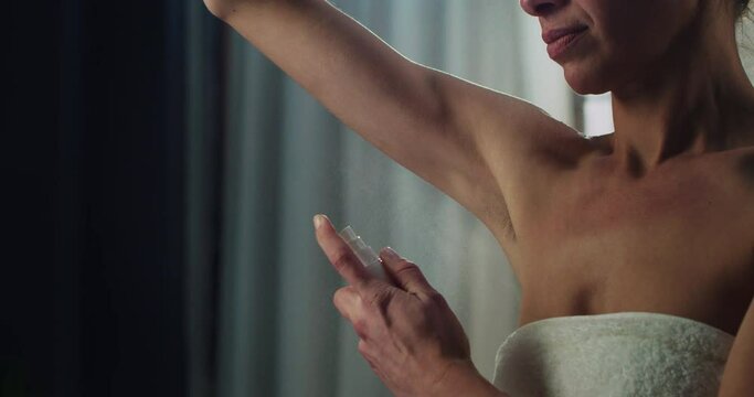 Cinematic close up of young woman in white bathrobe is applying female antiperspirant body spray deodorant on her armpit after shower during personal beauty hygiene routine at home before going out.