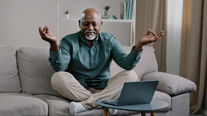 Calm mindful old 60s business man break sit at home on couch meditating breathing exercise feel no stress relief relaxing after work problem senior mature African male practicing yoga peaceful mind