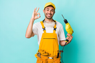 young adult hispanic man feeling happy, showing approval with okay gesture. worker and drill concept