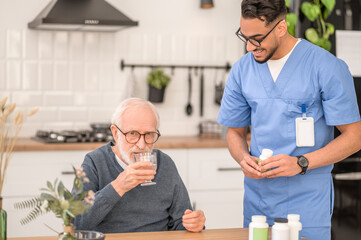 Young healthcare worker observing a pensioner swallowing down a medication - obrazy, fototapety, plakaty