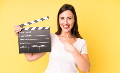 young adult pretty woman smiling cheerfully, feeling happy and pointing to the side. cinema clapper...