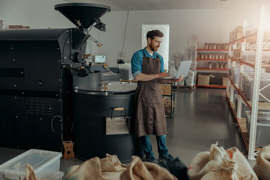 Business owner of coffee factory working laptop on background of coffee roasting machine - Powered by Adobe