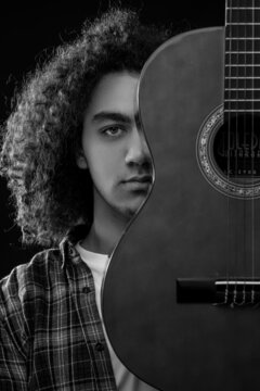 Young man with curly hair is posing with a acoustic guitar in front of his face. Black and white photo.