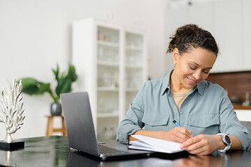 Woman is using laptop computer for remote work or studying online, she takes notes watching webinars or classes at kitchen table at home