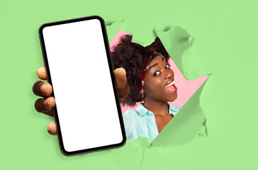 Happy excited young african american female with open mouth looks through hole in green paper and shows smartphone