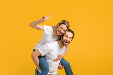 Smiling millennial european man and woman in white t-shirt take wife on back and have fun, lady...