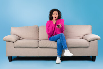 Frightened young African American lady with remote control sitting on sofa and watching scary movie or thriller on TV