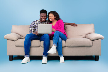 Happy young black couple using laptop while sitting on sofa over blue studio background, full length