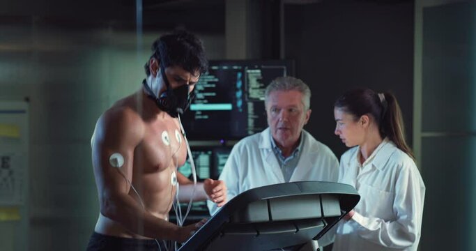Cinematic shot of young athlete with medical electrodes doing endurance test of body resistance by training on treadmill while physicians monitoring heart rate, pulse and ECG data in modern laboratory