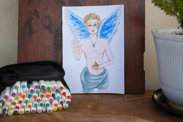 Handsome angel on paper and set of alcohol markers on wooden table at art studio. Hand drawn...