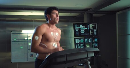 Cinematic shot of young athlete with medical electrodes doing endurance test of body resistance on treadmill and using futuristic screen for graphic implementation in modern laboratory.