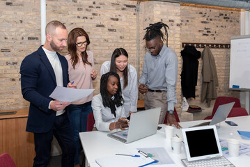 Young multi-ethnic business team working on a project, sharing ideas and analyzing data around a colleague sitting at the table looking at the computer in an office