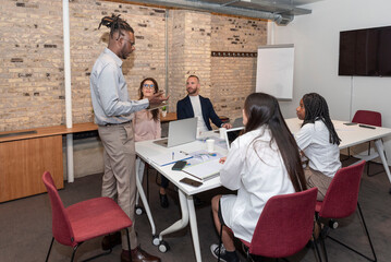 african american executive leader with a group of employees at the corporate coworking office meeting. Multiracial co-workers listening to boss explain new strategy plan at team meeting