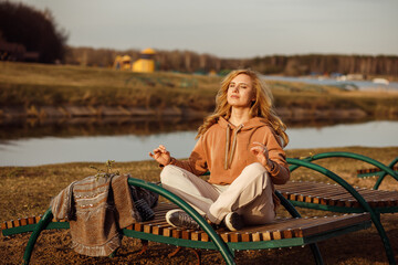 Beautiful blonde woman sitting alone on bench next to river bank on cool sunny day, relaxing, meditating and enjoying peace and freedom. Outdoors weekend activity, rural nature background