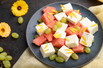 Vegetarian salad with watermelon, feta cheese, and grapes on blue ceramic plate on black concrete background. Side view, selective focus.