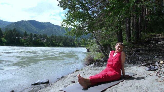 Old woman sitting on river bank on  yoga mat enjoying the sunny summer warmth, silence and tranquility