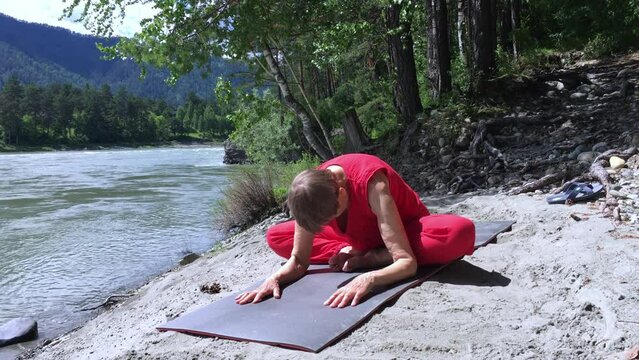 elderly woman yoga teacher sitting on flowing river shore doing the exercise bend to bent legs yoga asana