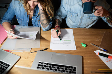 Crop of long-haired woman reading documents while man drinking coffee and underlining text. Couple in denim shirts working together at desk.