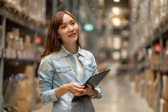 Asian female employee walks with a tablet to count inventory and look to buy raw materials in stock on a shelf in a warehouse.