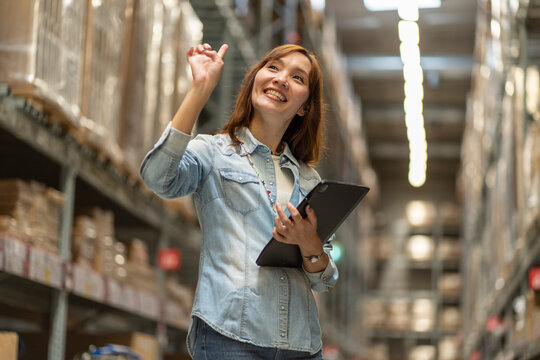Asian female employee walks with a tablet to count inventory and look to buy raw materials in stock on a shelf in a warehouse.