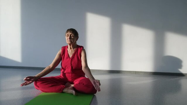 Old woman doing yoga sitting on  mat in white sunny studio meditating alone