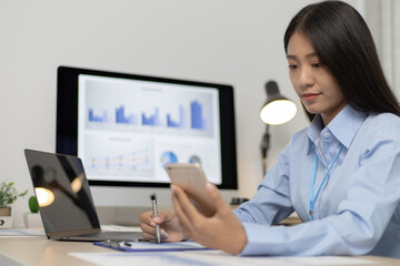 Asian female employees are analyzing market data via smartphones and have laptops to calculate the...