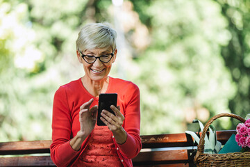 Gorgeous smiling caucasian senior woman with blonde short hair sitting on bench in park and make...