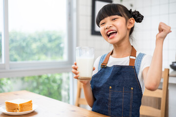 Asian little cute kid holding a cup of milk in kitchen in house. Young preschool child girl  stay home with smiling face, drinking milk and then look at camera.Milk for good health.