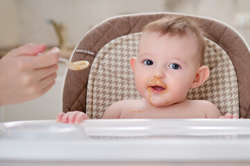 Mother feeding toddler baby from a spoon on a high chair for children, home kitchen background. Child boy at age of six months eats applesauce while sitting on a baby chair