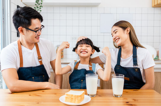 Asian  Family Enjoying Breakfast At Cozy Kitchen, Little Girl Daughter Sitting On Table, Drinking Milk With Smiling Father And Mother In Morning. Happy Family In Kitchen.