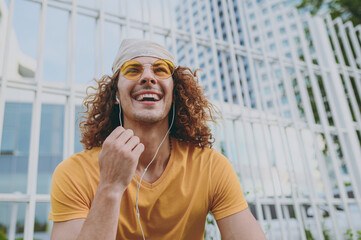 Bottom view young fun cool man 20s wearing yellow t-shirt bandana sitting listen to music in earphones headphones talk call rest relax in city town outdoors on nature. Urban lifestyle leisure concept