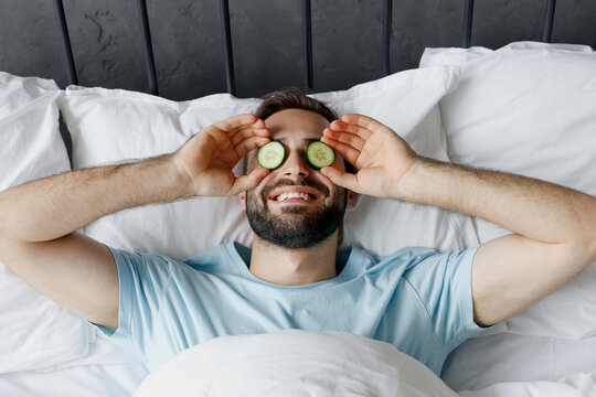 Young smiling fun man 30s wear casual blue t-shirt put cucumber slices on eyes rejuvenation mask lying in bed rest relax spend time in bedroom lounge home in own room house. Skin care routine concept.