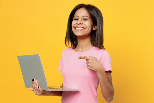 Little fun kid girl of African American ethnicity 12-13 years old in pink t-shirt hold use work point finger on on laptop pc computer isolated on plain yellow background. Childhood lifestyle concept.
