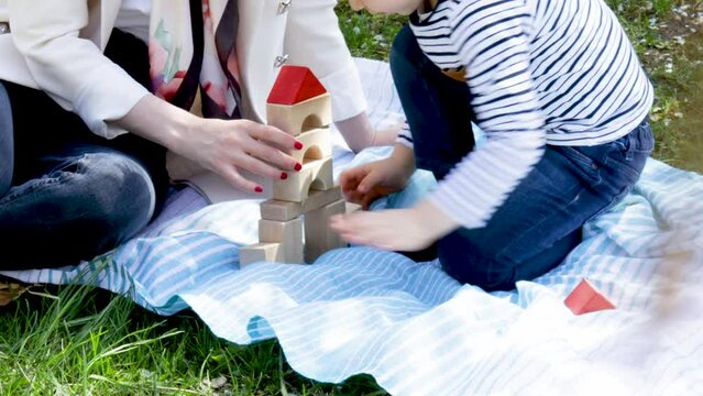 young mother and kids, little boy, is playing outdoors, in park, on blanket with eco wooden cubes. babysitter, woman, girl and child are building house or castles. kids activities, buy a house,rent
