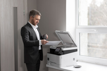 Adult successful man in a gray business suit standing near a copier with a cup of coffee