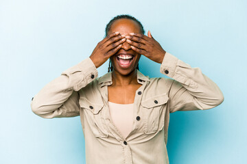 Young african american woman isolated on blue background covers eyes with hands, smiles broadly waiting for a surprise.