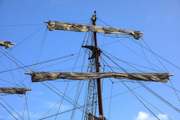 Masts, rigging and sails of an old historical sailing ship in a harbor against a blue sky with few...