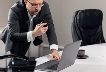 Handsome adult man in a gray suit holds a phone and takes pictures of his colleague's laptop