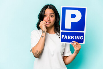Young hispanic woman holding parking placard isolated on blue background biting fingernails,...