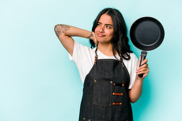 Young hispanic cooker woman holding frying pan isolated on blue background touching back of head,...