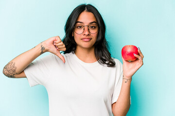 Young hispanic woman holding an apple isolated on blue background showing a dislike gesture, thumbs...
