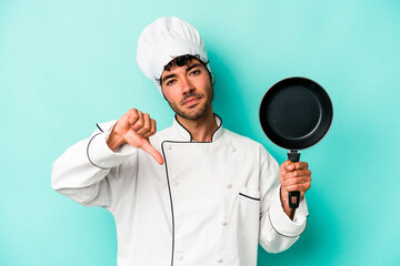 Young caucasian chef man holding flying pan isolated on blue background showing a dislike gesture, thumbs down. Disagreement concept.