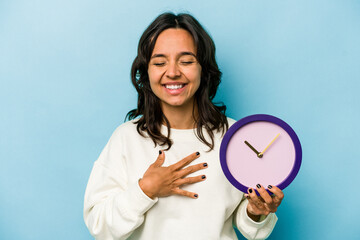 Young hispanic woman holding a clock isolated on blue background laughs out loudly keeping hand on...